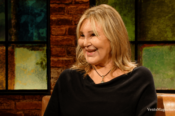 Carol Bryan smiling during a television interview, wearing a black top in a warmly lit studio setting with artistic background panels.