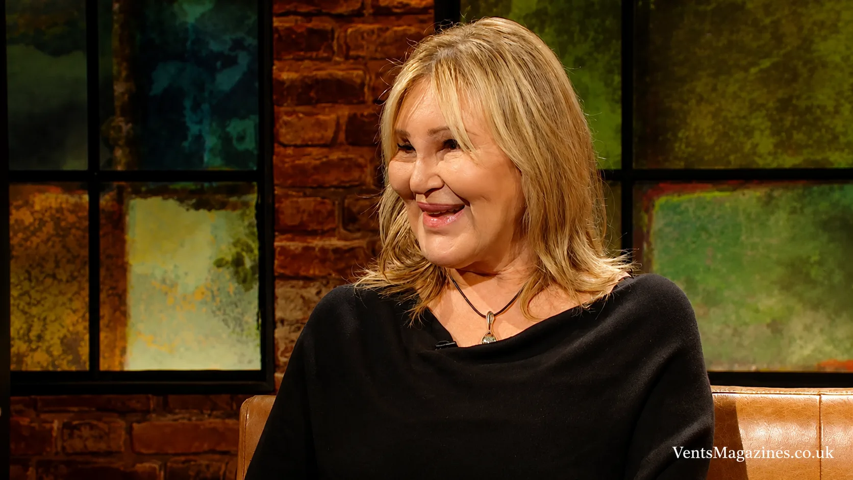 Carol Bryan smiling during a television interview, wearing a black top in a warmly lit studio setting with artistic background panels.